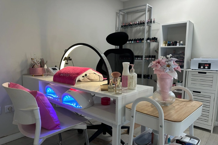 Einzelne Nagelstation mit einem weissen Tisch, einem Ringlicht und zwei blau leuchtenden LED-Lampen, einem schwarzen Stuhl und Regalen voller bunter Lacke, aufgelockert durch eine rosa Vase mit Blumen.