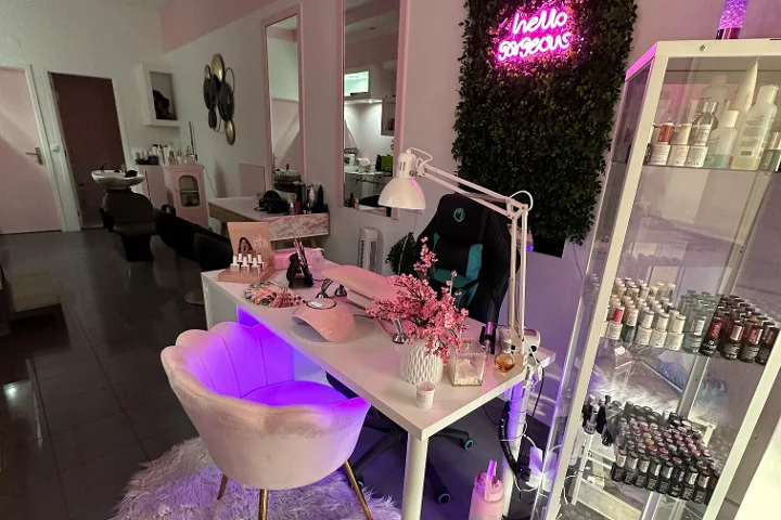 Rosa beleuchteter Beauty-Arbeitsplatz mit einem 'hello gorgeous'-Neonschild auf einer Grünwand, einem cremefarbenen Muschel-Sessel, einem weissen Tisch mit Pinseln und Kirschblüten, und einer Glasvitrine voller bunter Nagellacke.