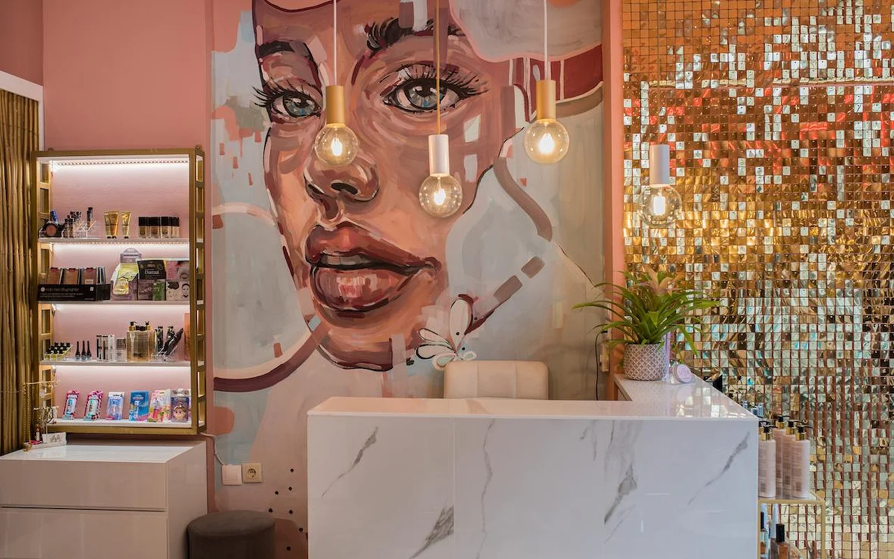 Ein Salon mit rosa Wänden mit einem grossen gemalten Wandbild eines Frauengesichts, einer schimmernden goldenen Paillettenwand, einer Rezeptionstheke aus weissem Marmor und Pendelleuchten mit freiliegenden Glühbirnen.