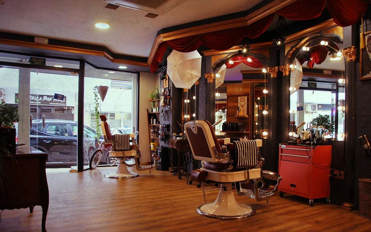 Ein Barbershop im Vintage-Stil mit klassischen Liegesesseln aus Leder auf verchromten Sockeln und gestreiften Handtüchern. Vergoldete Spiegel mit runden Glühlampen und roten Samtvorhängen säumen die Wand, ein roter Werkzeugwagen steht seitlich, und Fenster zur Strasse erhellen den Holzboden.