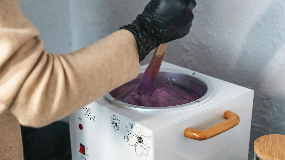 Die Hand einer Fachkraft in einem schwarzen Handschuh rührt in einem Kosmetikstudio in einem Topf mit geschmolzenem Warmwachs