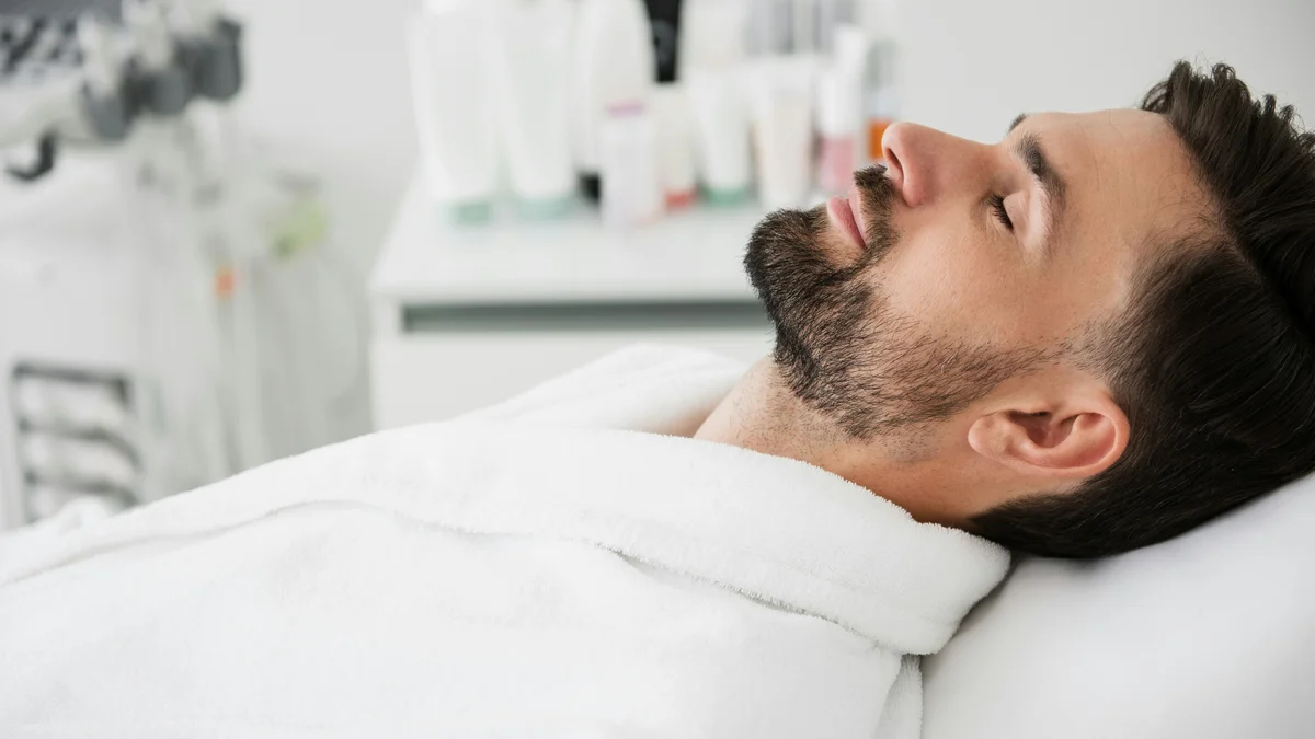 Ein Mann liegt in einem weissen Bademantel auf einer Behandlungsliege, den Kopf nach hinten gelegt und die Augen geschlossen