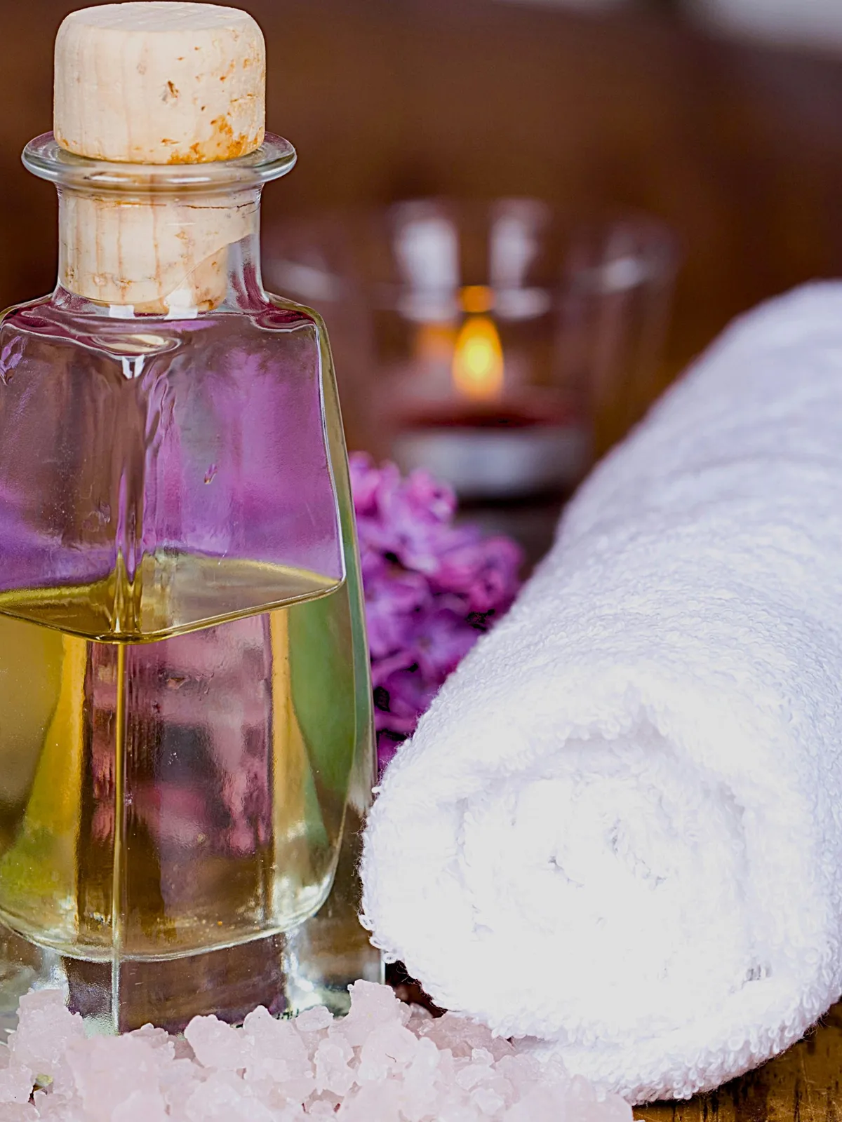 Nahaufnahme einer Glasflasche Öl mit Korken auf einem Tisch, daneben ein gerolltes weisses Handtuch und rosa Blüten