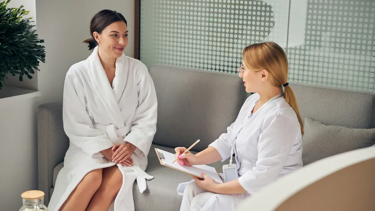 Eine Kundin im weissen Bademantel und eine Fachkraft mit Klemmbrett sitzen nebeneinander auf einem Sofa im Beautysalon, während die Fachkraft Fragen vor der Behandlung stellt und notiert